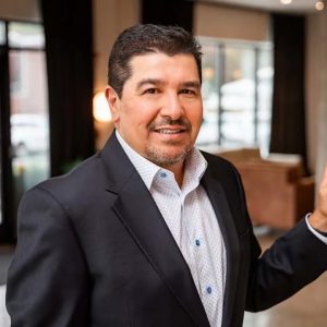 Man in a dark blazer and white dotted shirt smiles toward the camera in a modern indoor setting.