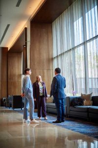 Three people in a modern lobby conversing by a large window with sheer curtains and blue seating area.
