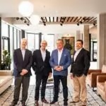 Five men in business attire pose in a modern office lobby with wooden beams and large windows.