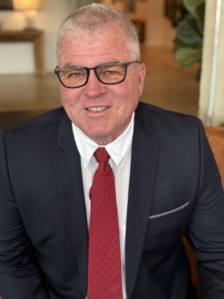 Burt Clark - Qualifind Group Smiling man in a dark suit, white shirt, red tie, and black-framed glasses, seated indoors.