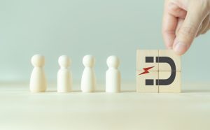 Line of four wooden figures as a hand places a block with a 'D' and red lightning bolt, symbolizing disruption in people/digital transformation.