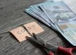 Scissors cutting a sticky note reading 'COST' beside a stack of cash on a wooden table, implying cost cutting.