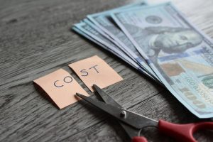 Scissors cutting a sticky note reading 'COST' beside a stack of cash on a wooden table, implying cost cutting.