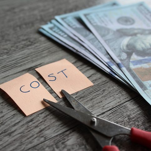 Scissor money and note with text COST Scissors cutting a sticky note reading 'COST' beside a stack of cash on a wooden table, implying cost cutting.