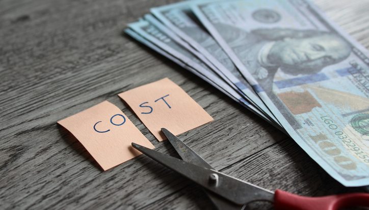 Scissors cutting a sticky note reading 'COST' beside a stack of cash on a wooden table, implying cost cutting.