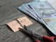 Scissors cutting a sticky note reading 'COST' beside a stack of cash on a wooden table, implying cost cutting.