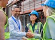 Diverse construction team shakes hands on a site with scaffolding behind them.