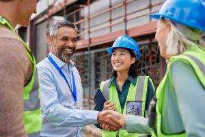 Diverse construction team shakes hands on a site with scaffolding behind them.