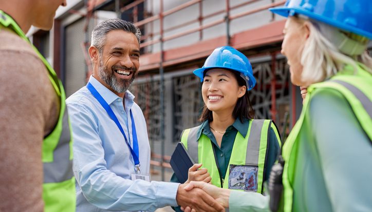 Diverse construction team shakes hands on a site with scaffolding behind them.