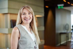 Xochilt Acosta - Qualifind Group Smiling blonde woman in a gray sleeveless vest and white blouse, standing in a modern office lobby.