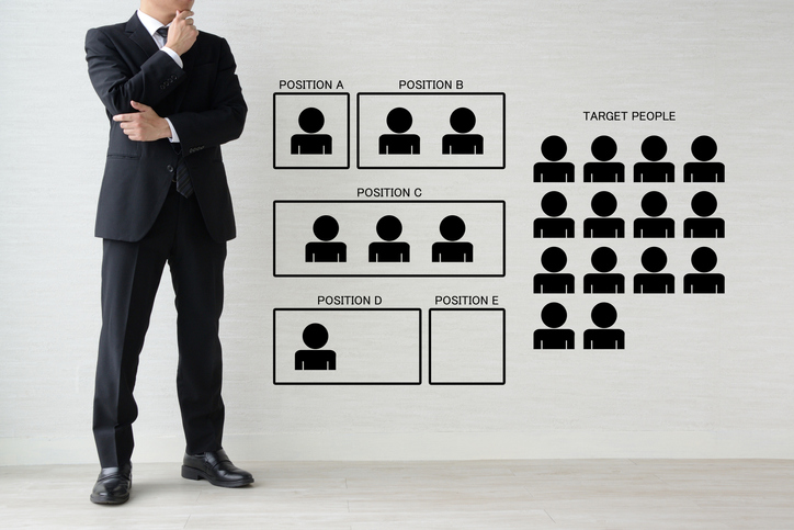 Trade Compliance Is No Longer a Back-Office Function Businessman in suit standing beside a wall chart showing recruitment positions A to E and a group of target people icons