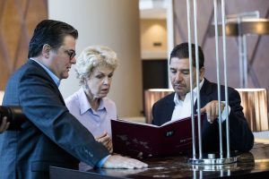 Three professionals in business attire review a red folder at a marble counter in a modern lobby.