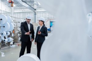 Two professionals in suits and hard hats discuss production at a textile factory with large white rolls nearby.